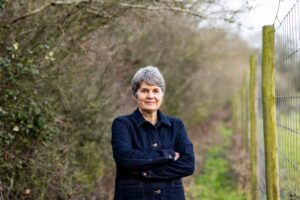 Sarah Finch standing with arms crossed on a footpath between a thick hedge and a wire fence
